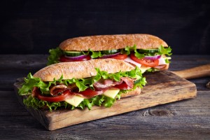 Zwei Sandwiches mit Schinken, Käse, Bacon, Tomaten, Salat, Gurken und Zwiebeln auf einem Holzbrett
