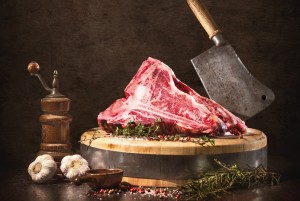 Rohes Dry Aged T-Bone Steak auf einem Holzbrett mit einem Beil und diversen Gewürzen